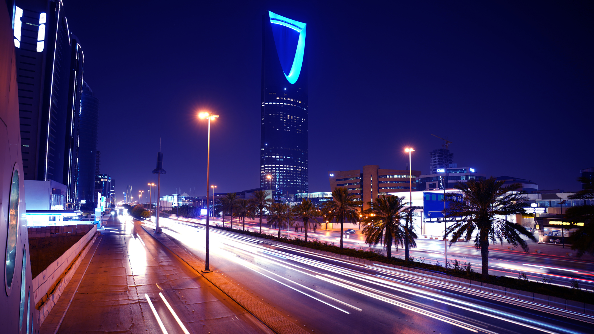 The Kingdom Centre Tower at night in Riyadh, seen from the highway with palm trees and car light trails, illustrating the urban life for Expatriates in Riyadh.