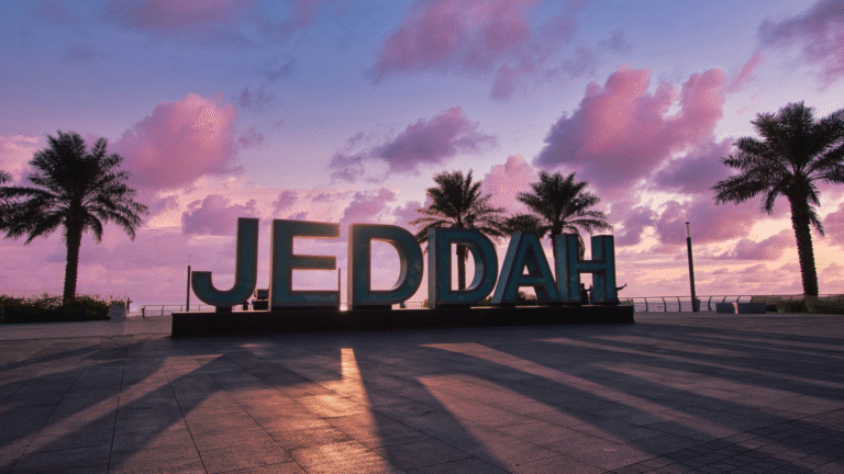 Iconic Jeddah sign at sunset with palm trees and purple sky, welcoming expatriates in Jeddah to Saudi Arabia's cosmopolitan coastal city.