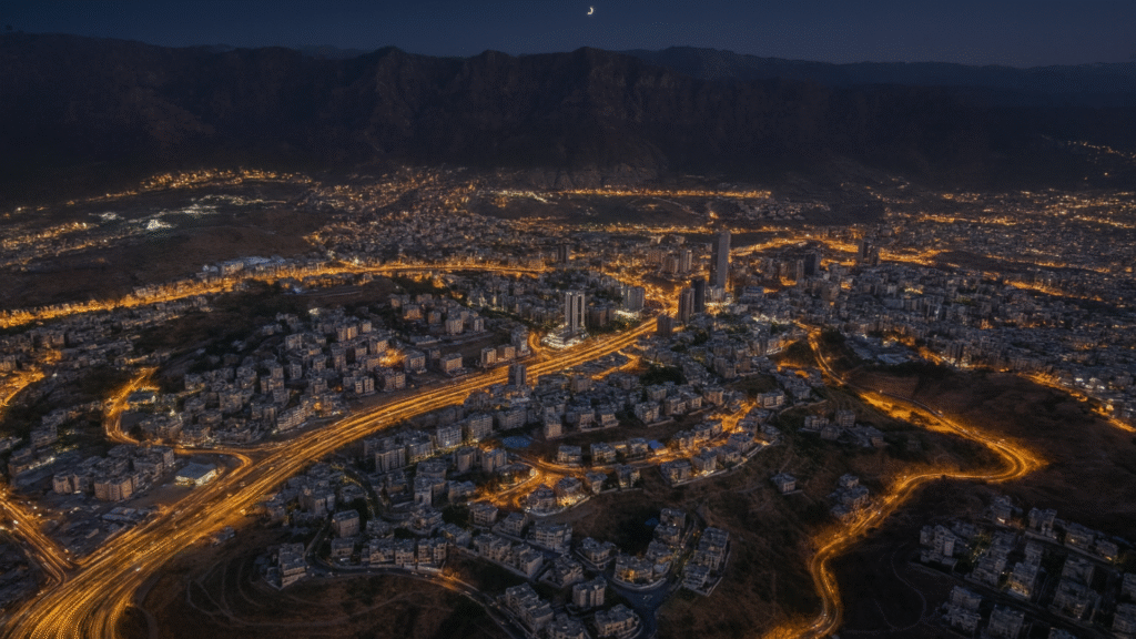 Realistic aerial night view of Khamis Mushait with illuminated roads, high-rise buildings, and surrounding mountains under a crescent moon – reflecting the life of expatriates in the region.
