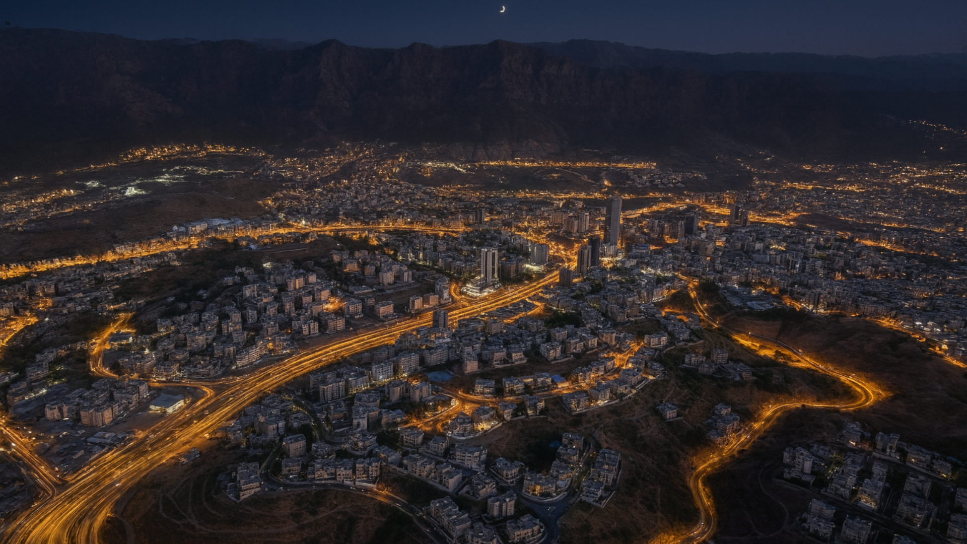 Realistic aerial night view of Khamis Mushait with illuminated roads, high-rise buildings, and surrounding mountains under a crescent moon – reflecting the life of expatriates in the region.